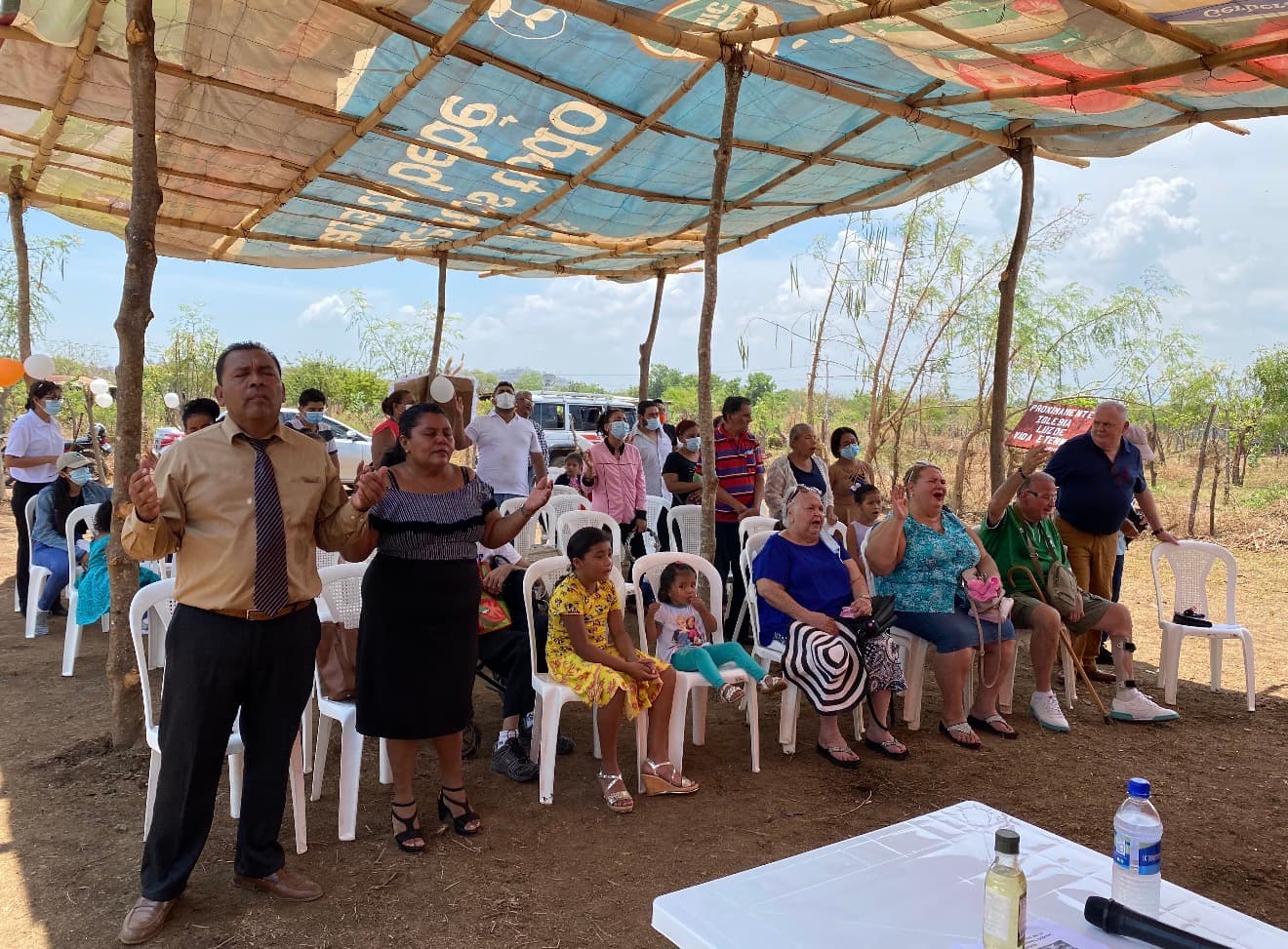 Hombre y mujer orando con manos alzadas bajo un toldo durante un servicio religioso comunitario.