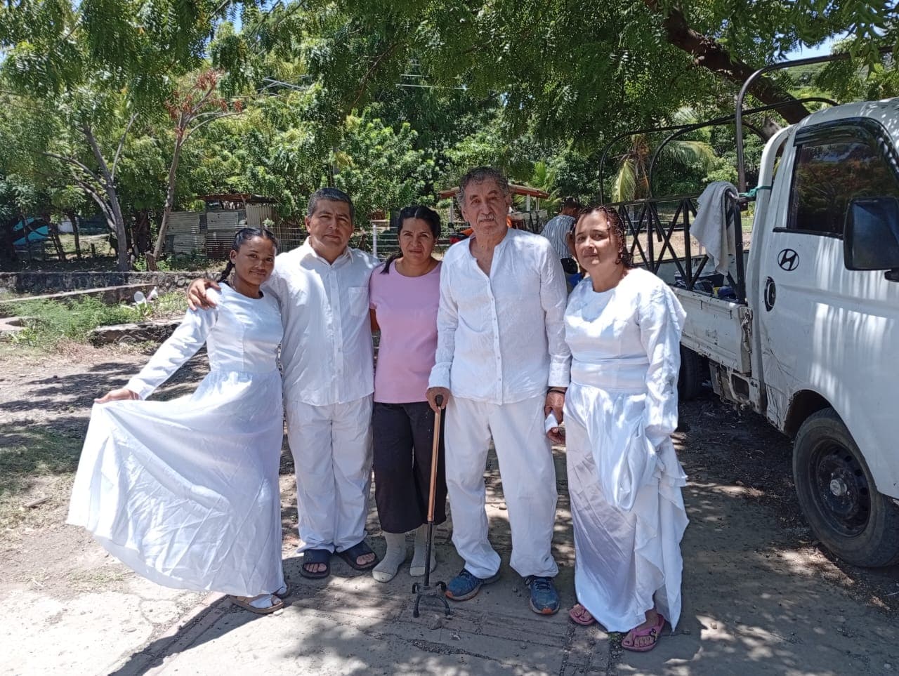 Cinco personas posan juntas, cuatro vestidas de blanco impecable bajo la sombra de árboles.
